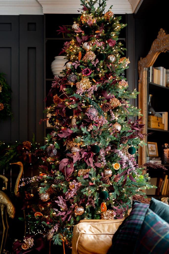 A beautifully decorated Christmas tree adorned with Christmas decorations in purple, brown, gold, and green ornaments, including flowers, baubles, and ribbons. The tree is set against a dark wall with a bookshelf and an ornate mirror nearby. Warm lights add a cozy glow.