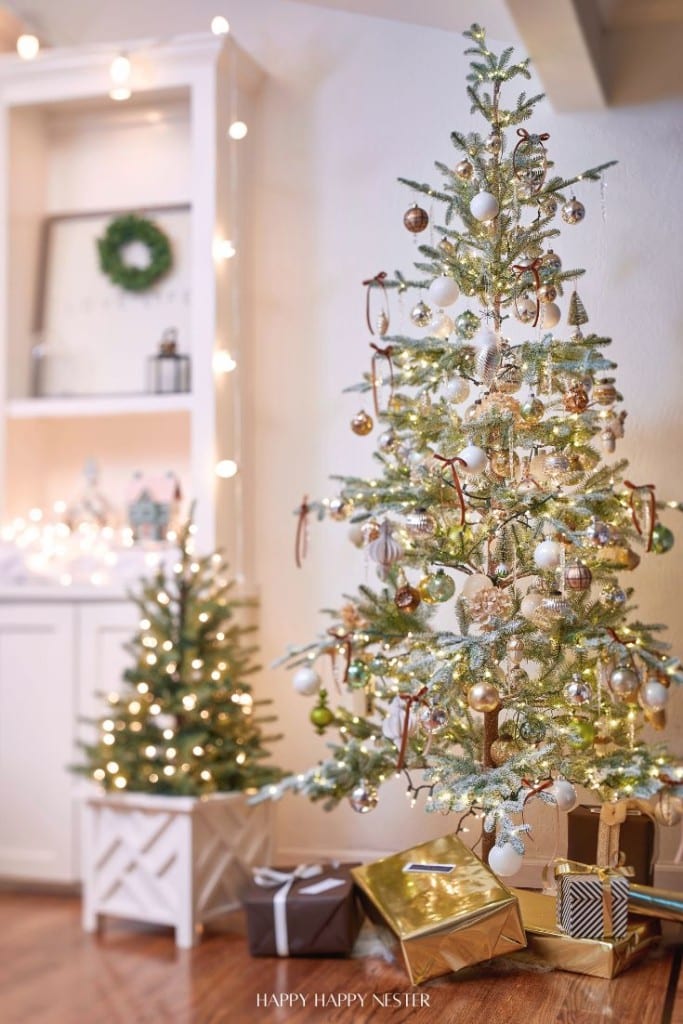 A festive room showcases a large Christmas tree with white and gold Christmas decorations and elegant ribbons. A smaller tree nearby glows with warm lights. The background displays a shelf of decorative items, while wrapped gifts rest at the base of the dazzling trees.