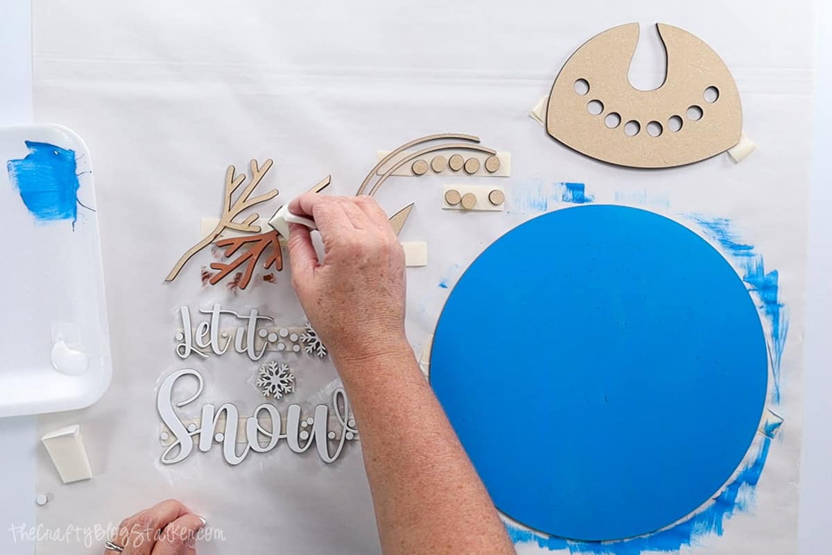 Painting in progress for a "Let It Snow" wood sign, with a blue background and detailed pieces like branches and text being painted brown and white for assembly.