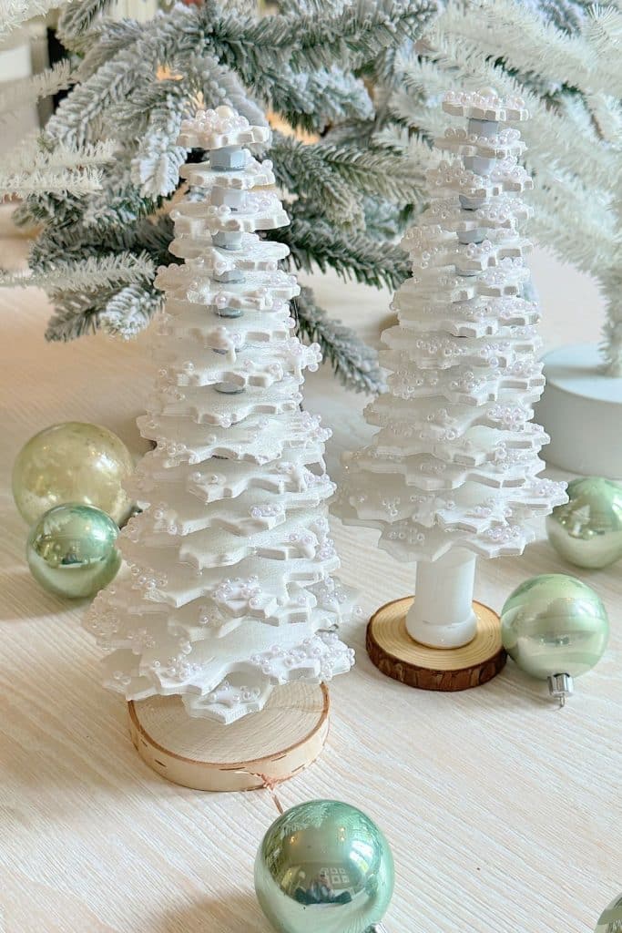 Two decorative white Christmas trees, crafted from air dry clay and adorned with small beads, sit on wooden bases. They are surrounded by shiny green ornaments on a light-colored surface, with snowy branches in the background.