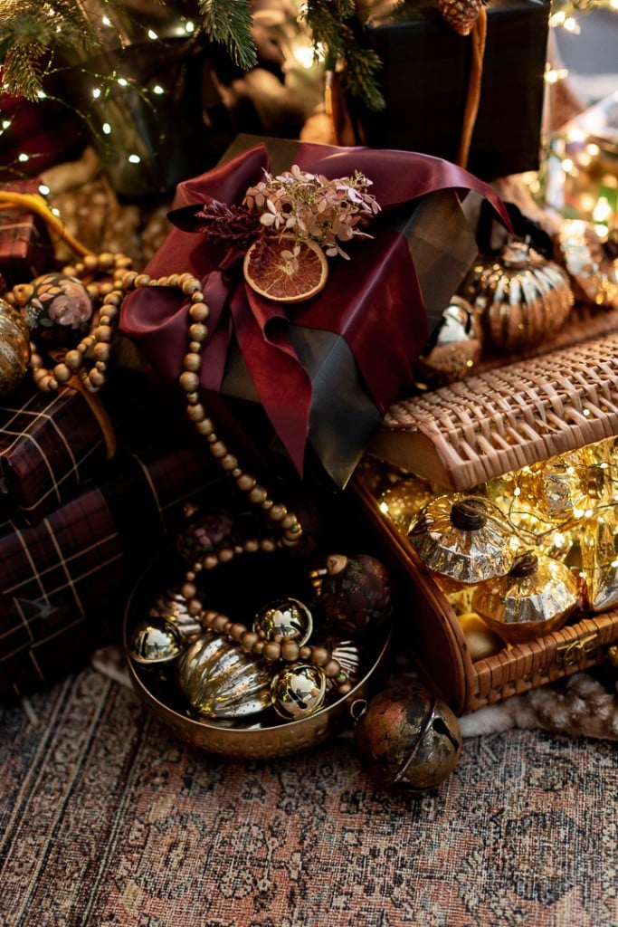 A festive Christmas display features wrapped gifts, gold ornaments, a woven basket of decorative balls, wooden beads, and a red napkin adorned with dried flowers and an orange slice. An air-dry clay house adds a charming touch, set elegantly on the patterned carpet.