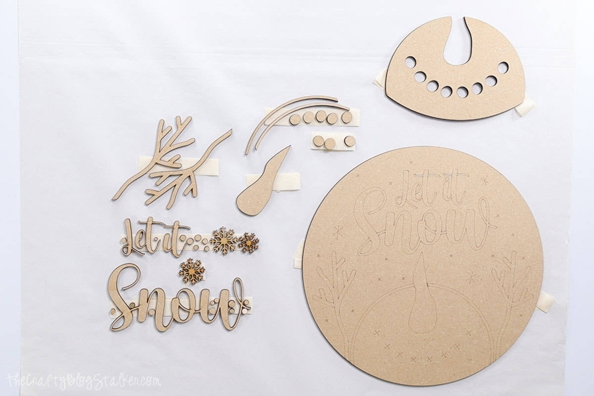 Laser-cut wood pieces for a "Let It Snow" sign, including text, snowflakes, branches, and snowman elements, laid out and ready for assembly and decorating.